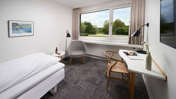 A hotel room with a bed, a desk with a chair, a window with a view of greenery, and a framed picture on the wall.