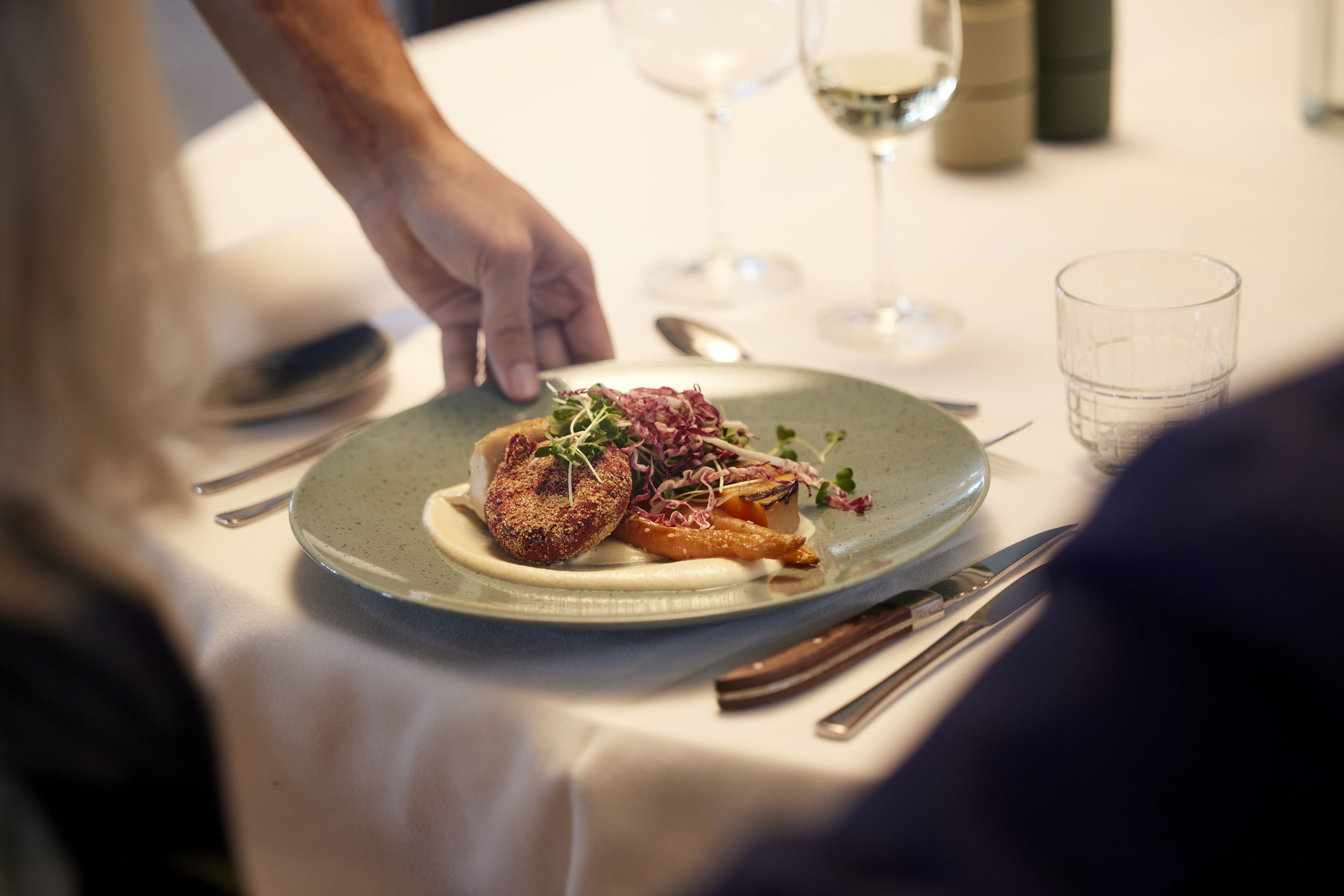 A plate of food with a hand reaching for it, a wine glass, and cutlery on a table.