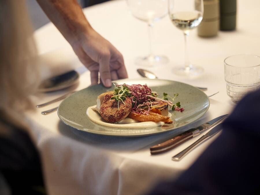 A plate of food with a hand reaching for it, a wine glass, and cutlery on a table.