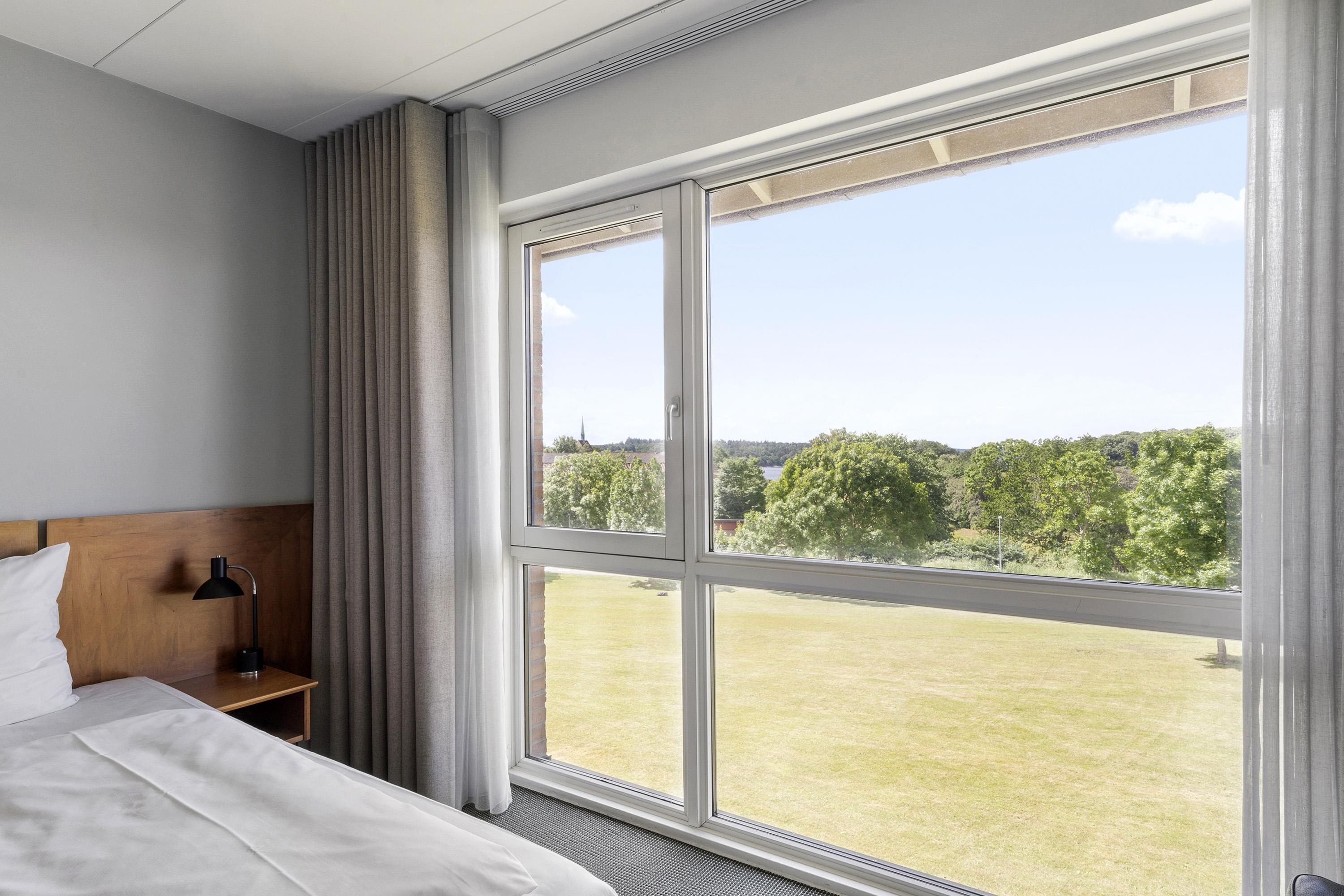 Quarto de hotel com uma grande janela, uma cama, uma mesinha de cabeceira e um abajur.