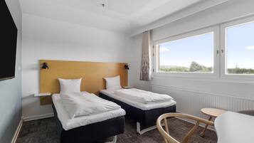 A hotel room with two single beds, a small table, a chair, and a large window with a view.