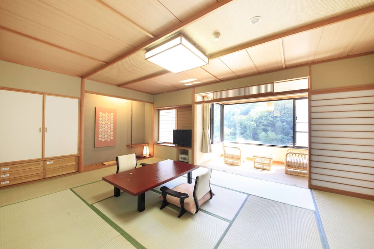 A traditional Japanese room with tatami flooring, a low table, chairs, a television, and a view of greenery outside.