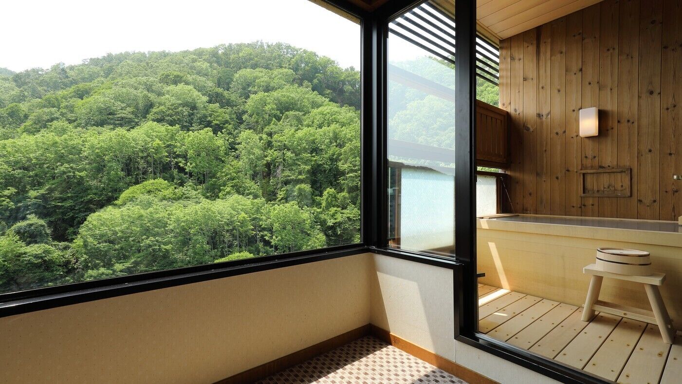 A room with a large window overlooking a forested area, a wooden deck, and a small stool.