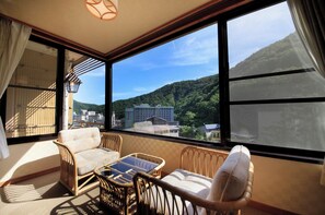 Un balcón con mobiliario de mimbre, vista a un paisaje montañoso y un edificio a lo lejos.