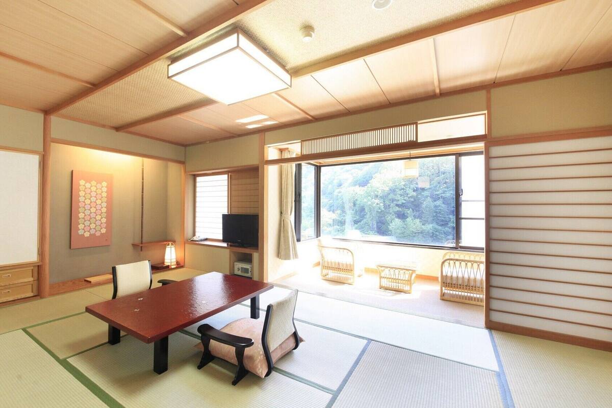 A traditional Japanese room with tatami flooring, a low table, chairs, a television, and a large window with a view of trees.