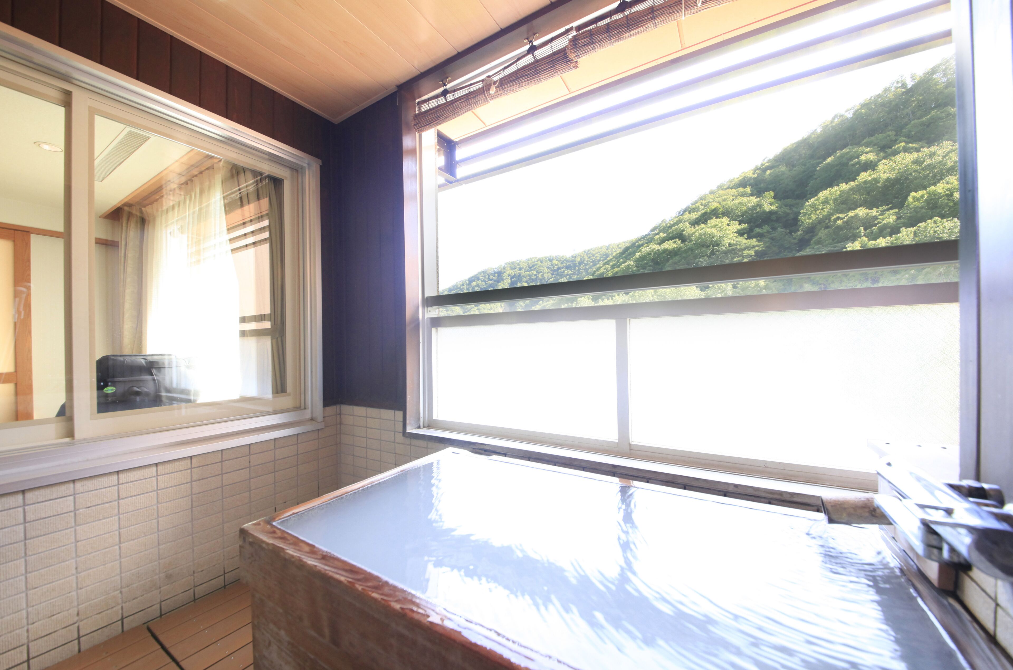 A bathroom with a bathtub, a window with a view of a forested hill, and a tiled floor.