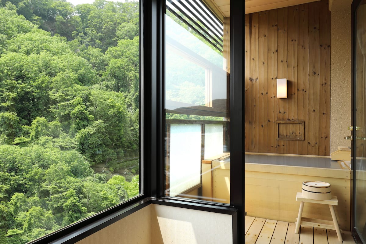 A room with a large window overlooking a forest, wooden walls, a small stool, and a built-in shelf.