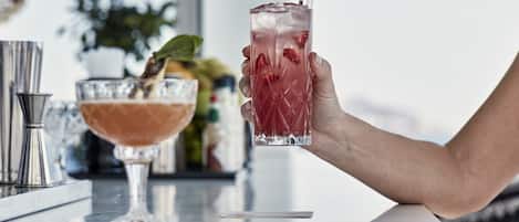 A hand holding a tall glass with a red drink and a straw, next to a shorter glass with a different drink.
