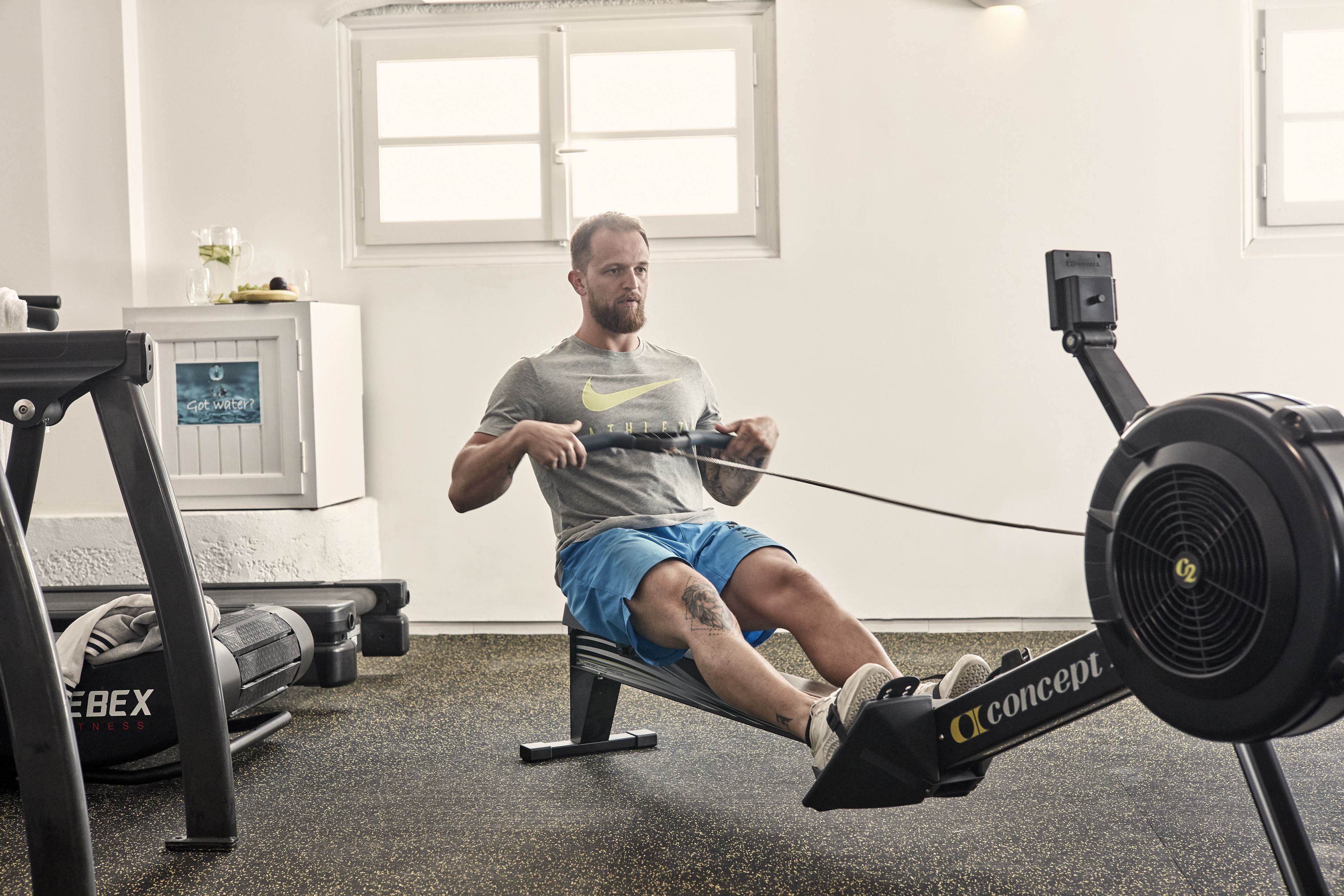 A man using a rowing machine in a gym with a window and a water dispenser in the background.