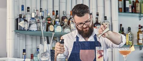 A bartender preparing a drink at a bar with shelves of liquor bottles in the background.
