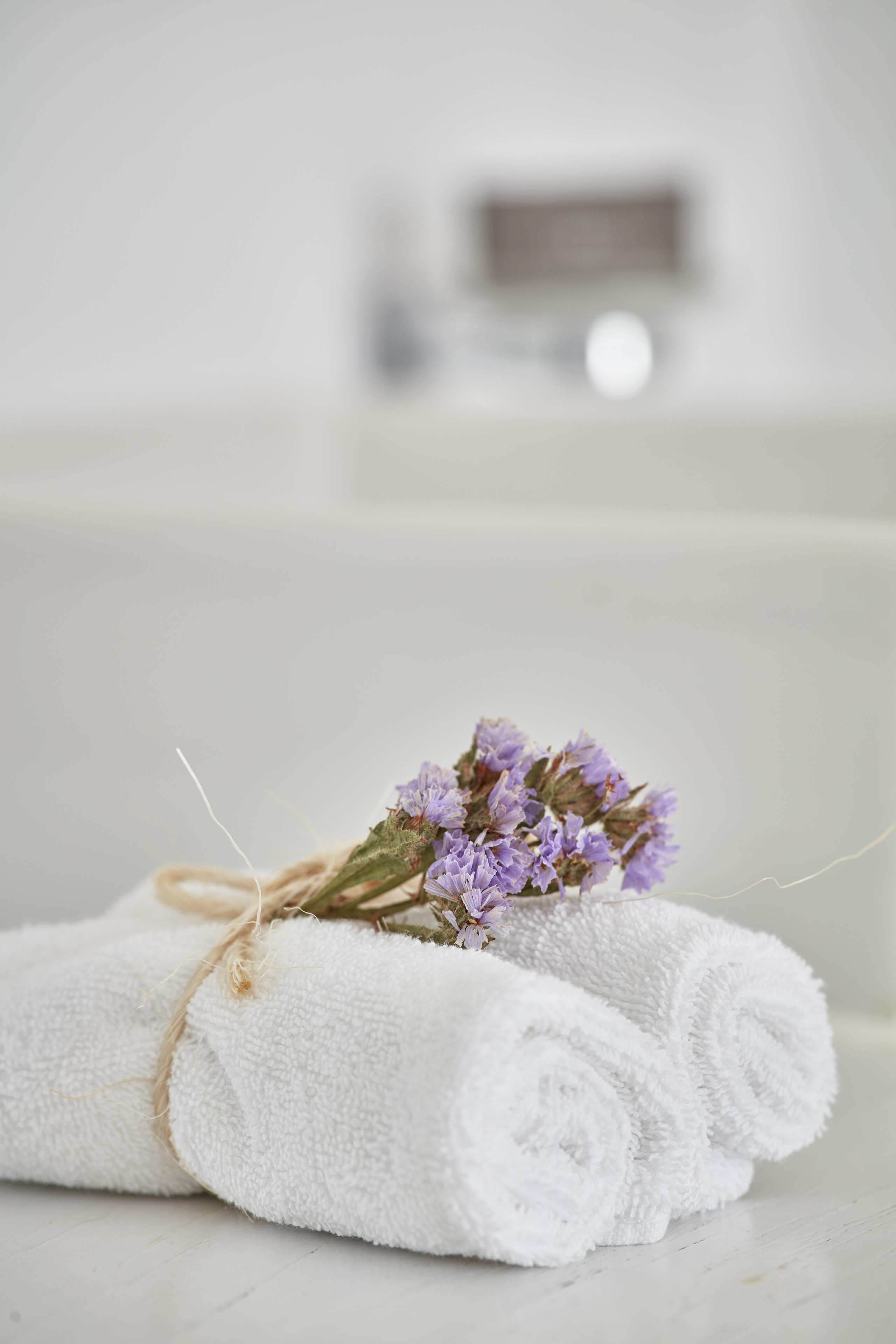 A white towel bundle with a small bunch of purple flowers tied with twine, placed on a white surface.