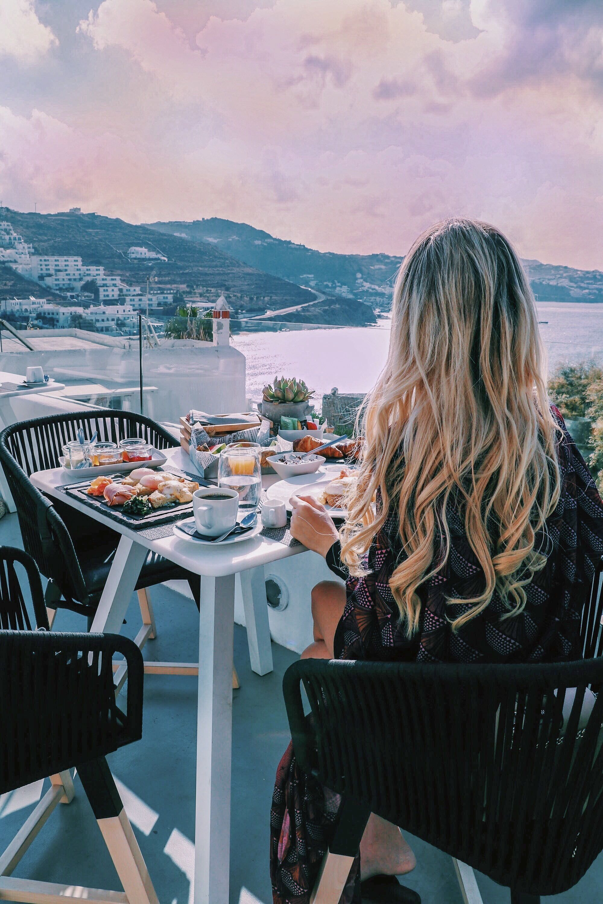 A dining table with food and drinks, overlooking a scenic view of a body of water and hills.