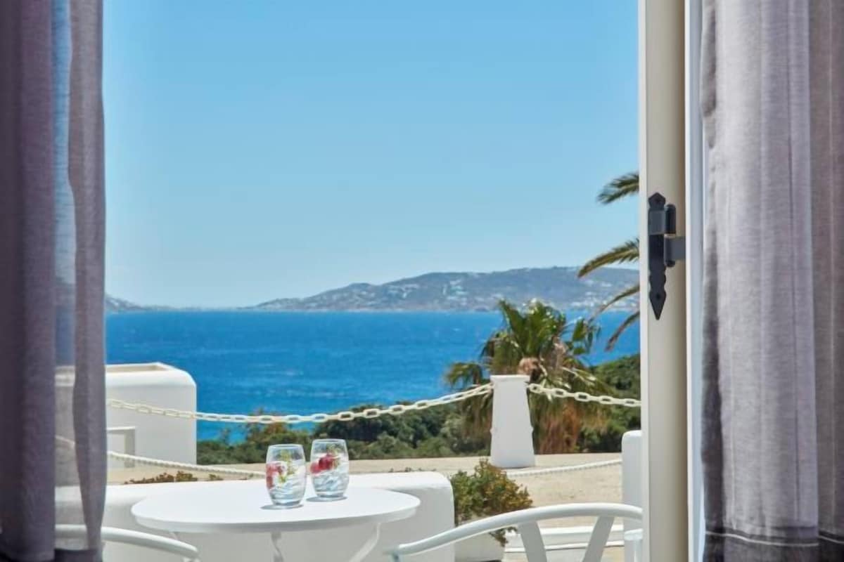 Un balcón con una mesa, dos vasos y vistas al mar y las colinas.