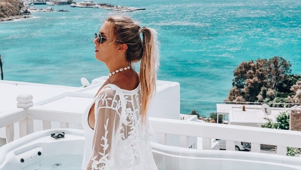 A person in a white, semi-transparent top sitting in a hot tub overlooking a coastal landscape. - Mykonos Princess Hotel (Mykonos)
