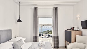 A modern hotel room with a large bed, a sitting area with a chair, a TV, and a balcony with a view of the sea. - Mykonos Princess Hotel (Mykonos)