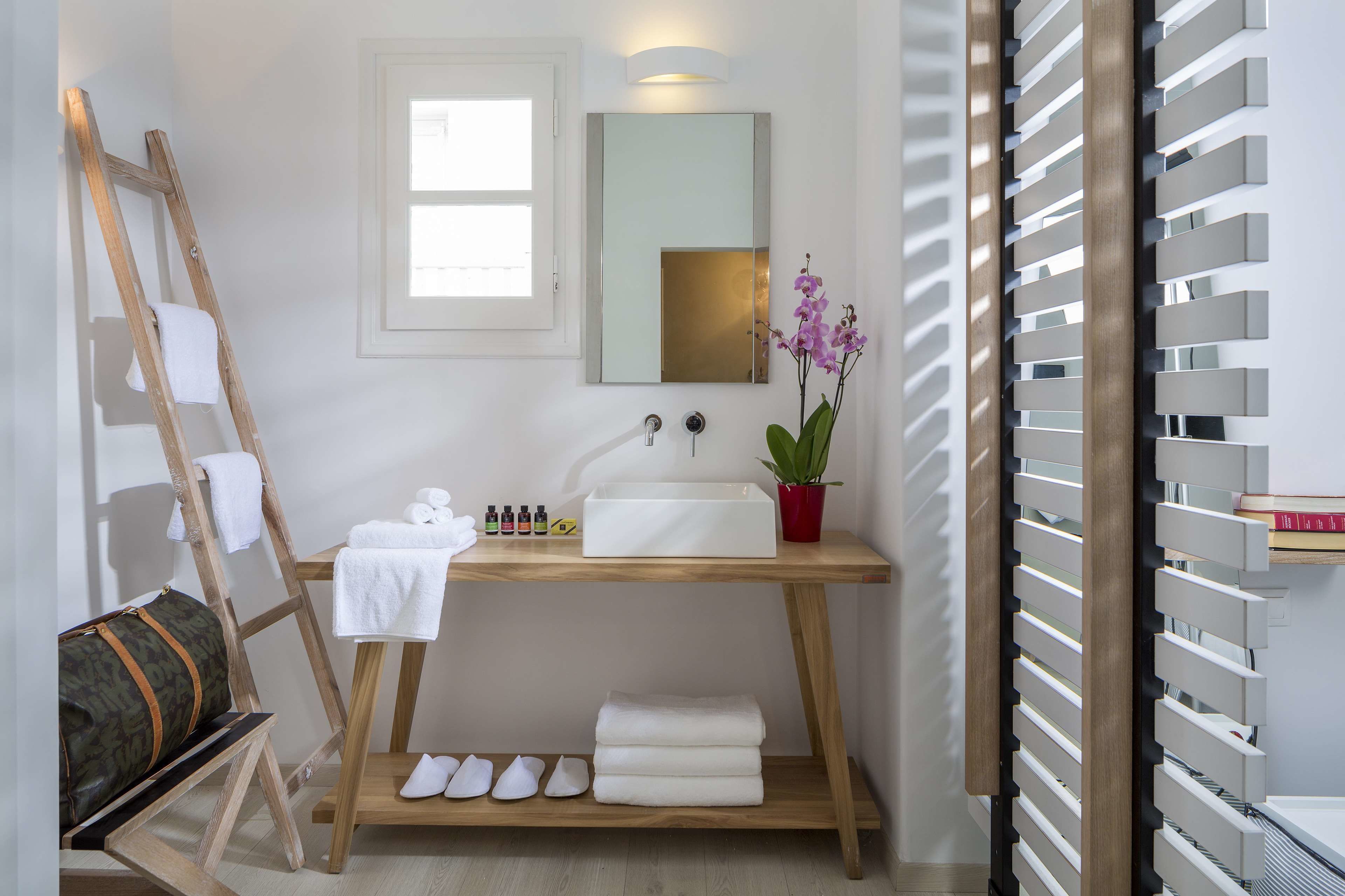 A bathroom with a wooden vanity, a mirror, a sink, a towel rack, a ladder, a potted orchid, and a shelf with books.