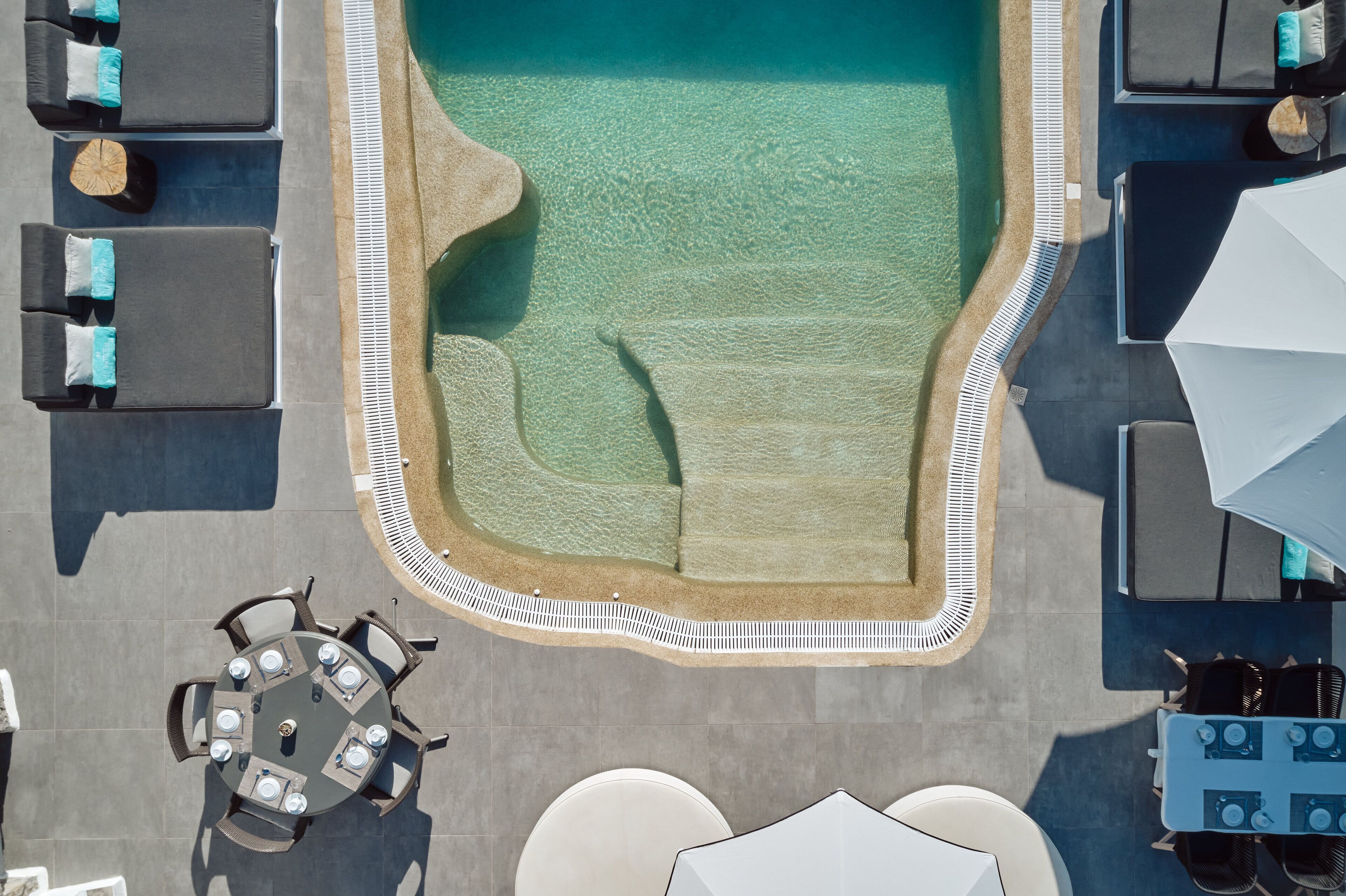 Vista aérea de uma área de piscina com espreguiçadeiras, mesa de jantar e guarda-sóis.