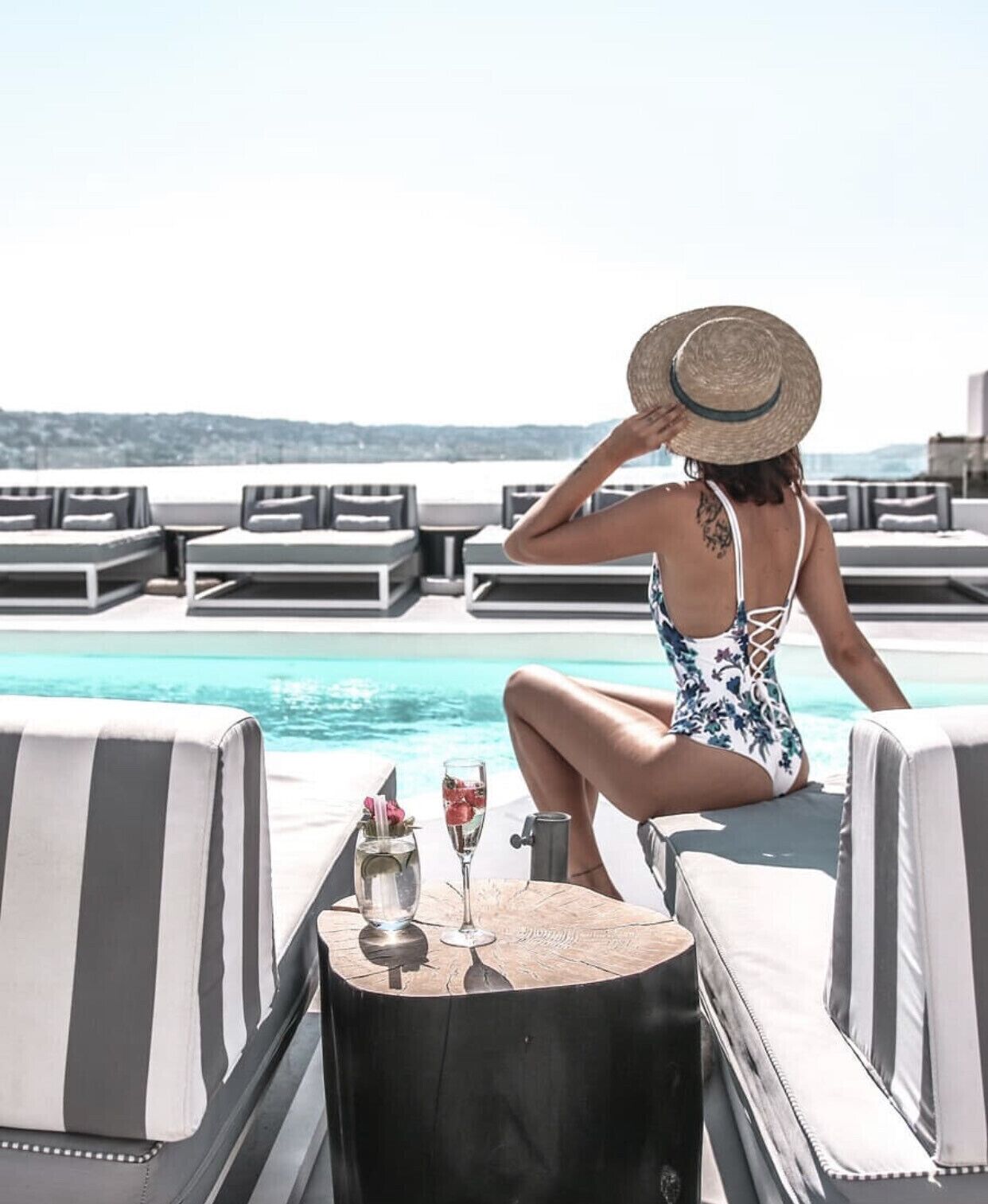 A woman sitting by a poolside lounge chair with a drink and a glass of water on a small table.