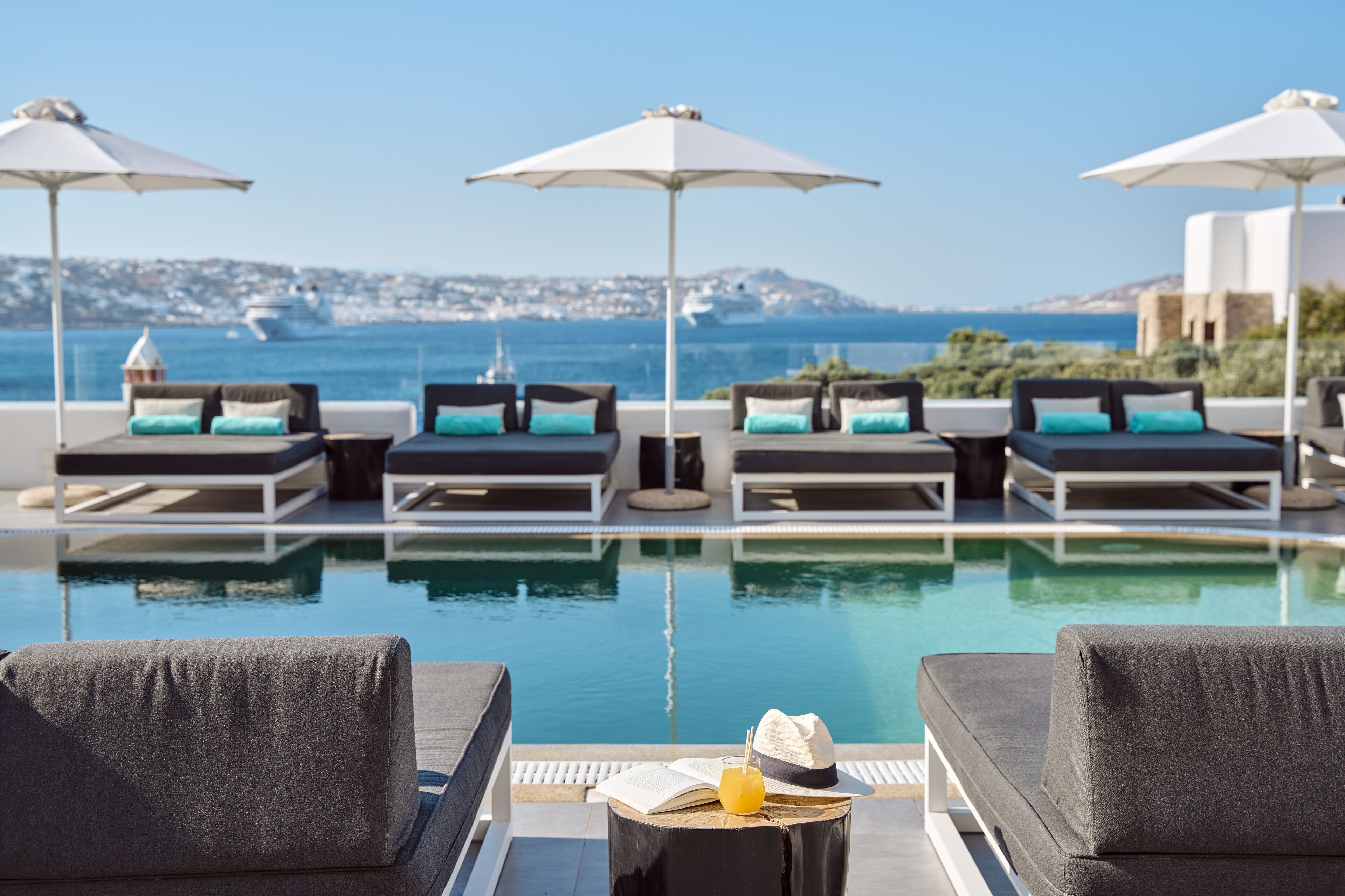 A poolside area with lounge chairs and umbrellas, a book, and a drink on a table, overlooking a body of water and distant land.