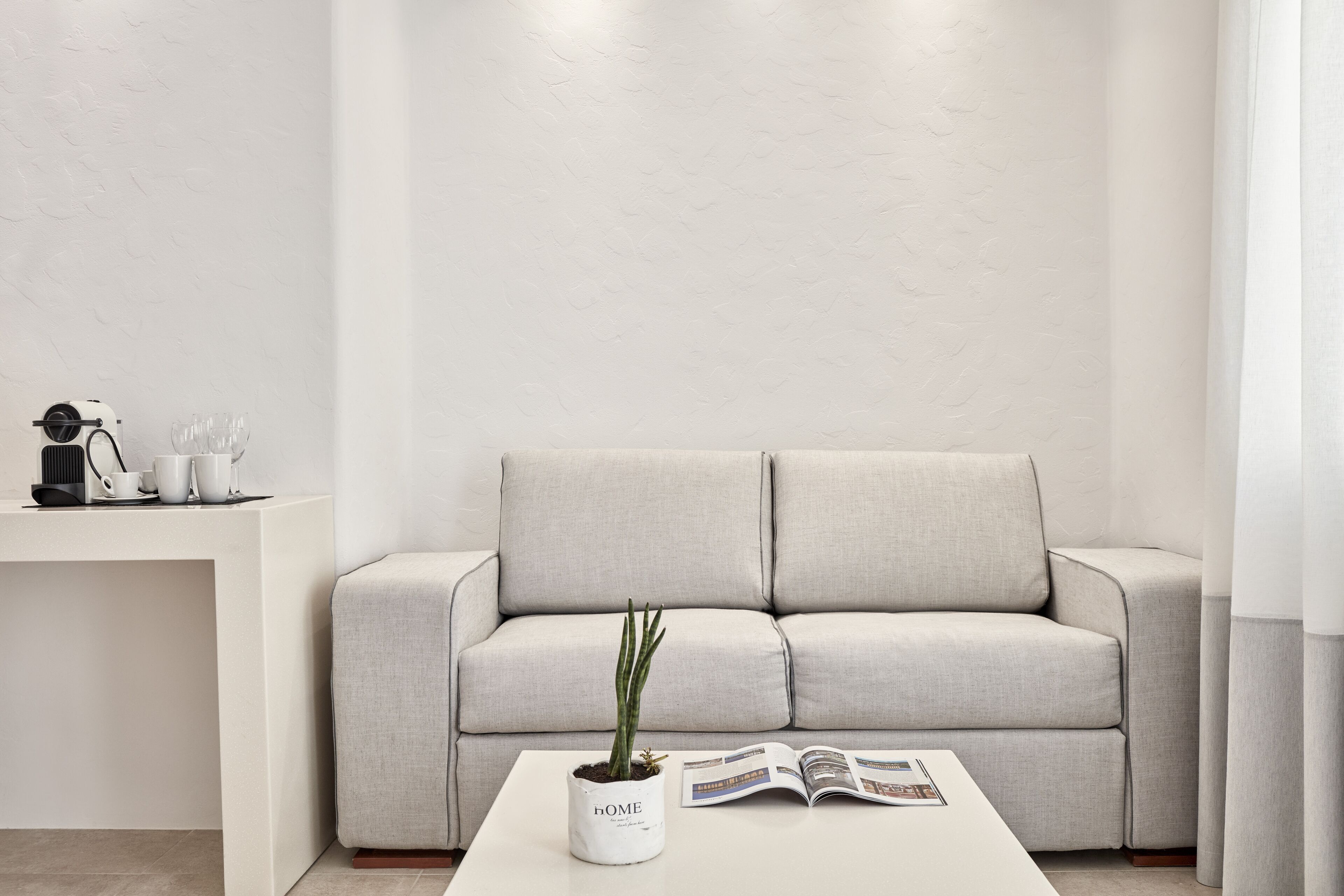 A minimalist living room with a light grey sofa, a white coffee table with a potted plant, and a magazine.