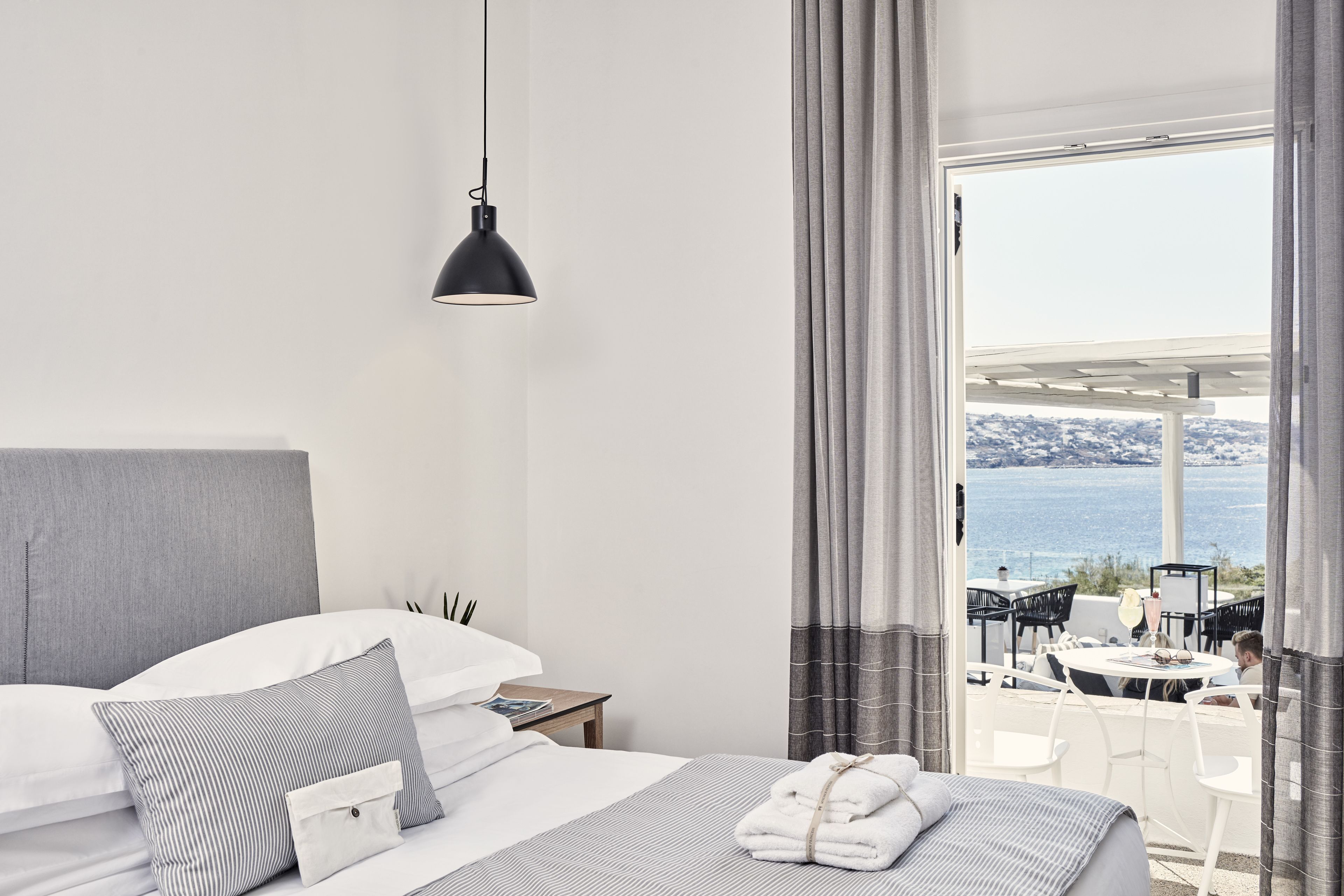 Une chambre d’hôtel avec un lit, une table de chevet, une lampe et une vue sur la mer grâce à de grandes fenêtres.