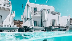 A swimming pool with a person swimming, surrounded by white buildings with balconies and outdoor seating. - Mykonos Princess Hotel (Mykonos)