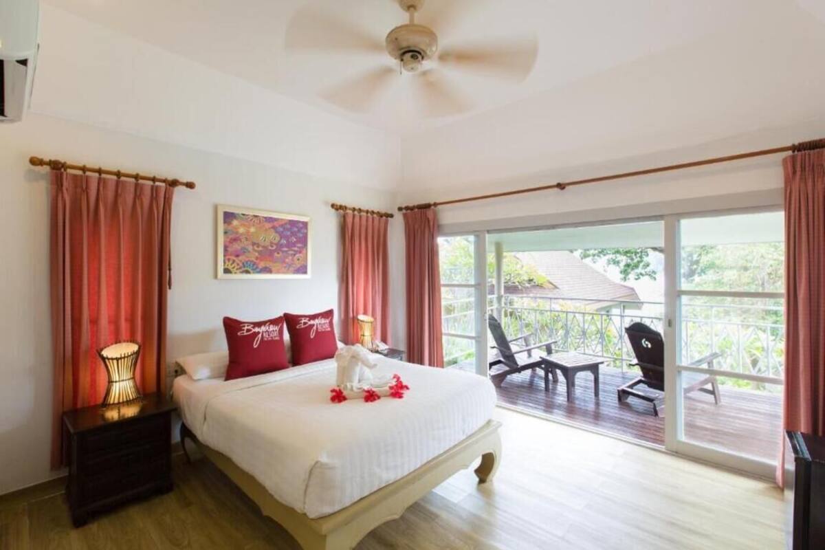 Une chambre avec un grand lit, un ventilateur de plafond, une table de chevet avec une lampe et un balcon avec des chaises.