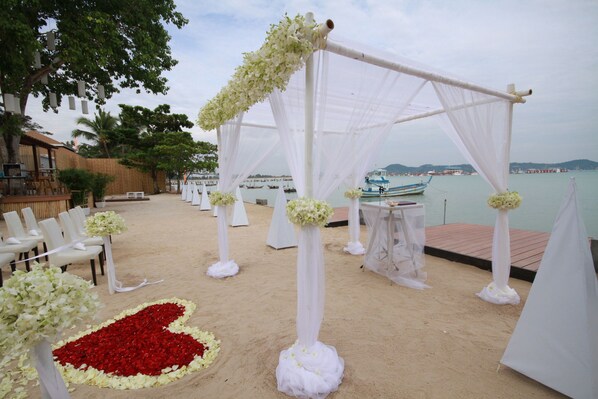 Decoración de boda al aire libre con suelo de arena, adornos florales blancos y vista a barcos en el agua.