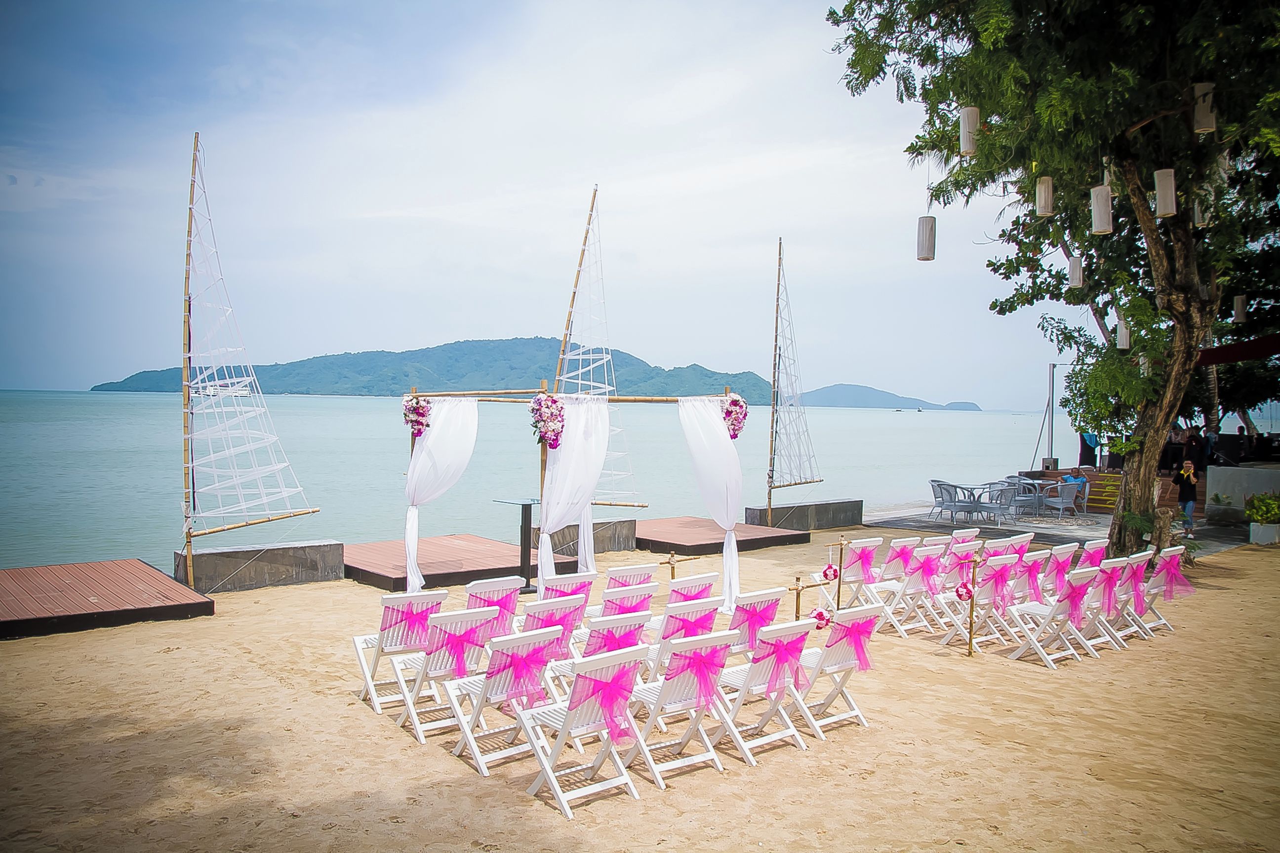 En bröllopsplats på stranden med vita och rosa stolar, en scen med vita draperier och en bakgrund bestående av vatten och kullar.