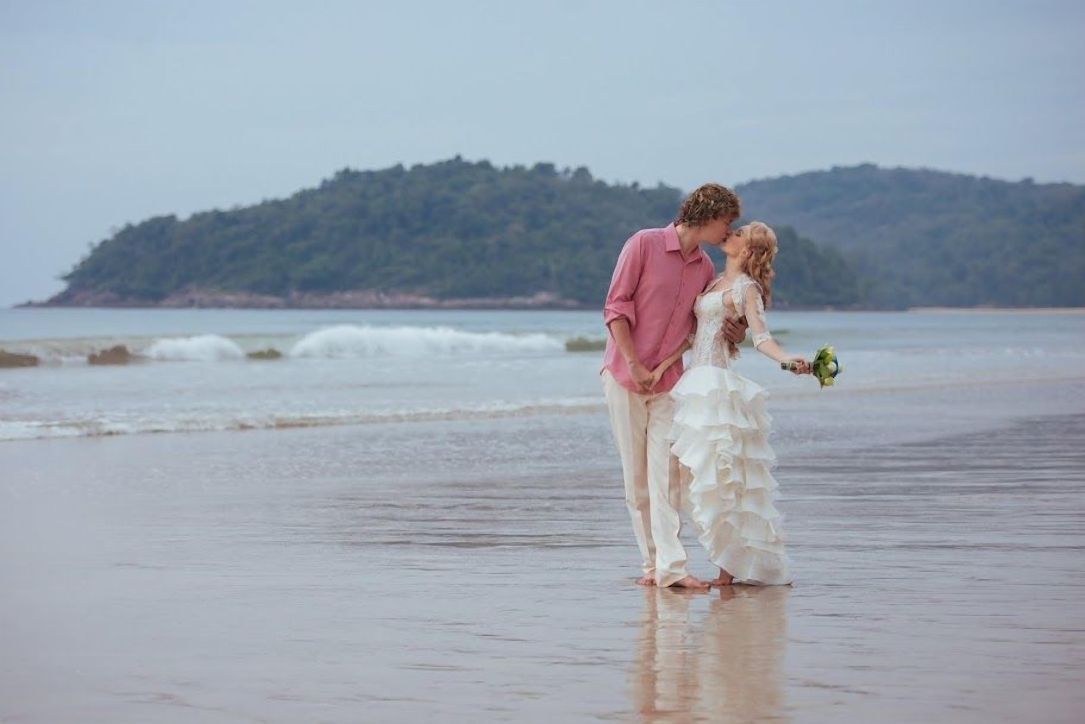 A couple standing on a beach with a view of the sea and a hill in the background.