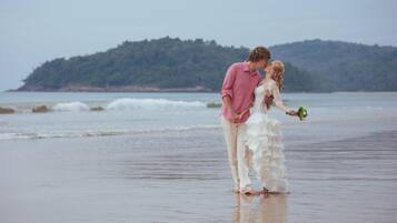 A couple standing on a beach with a view of the sea and a hill in the background.