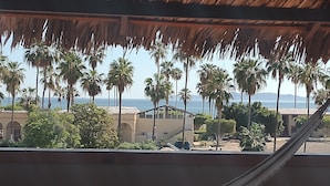 Una hamaca bajo un techo de paja, palmeras y un edificio con ventanas de arco.