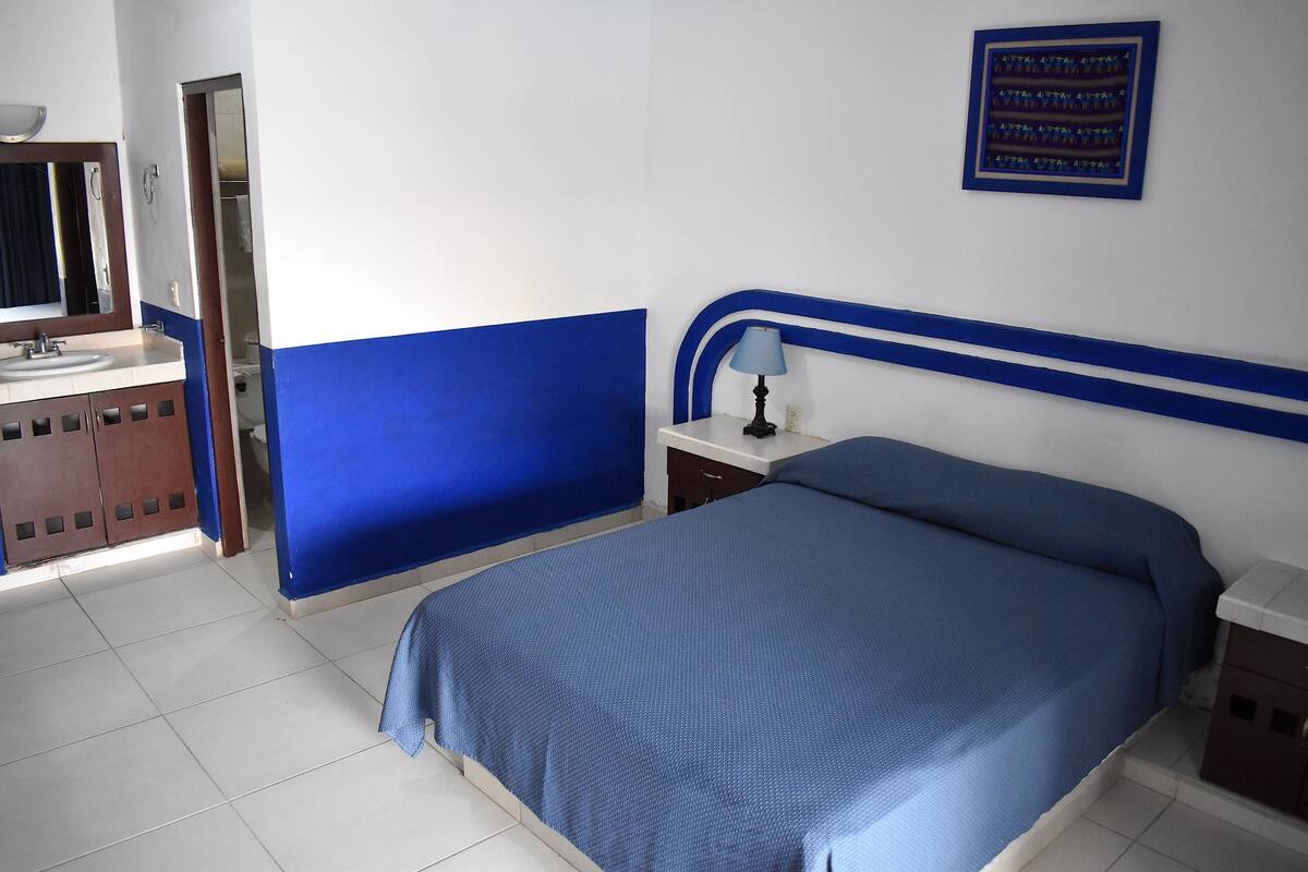 A bedroom with a bed, a nightstand, a lamp, a mirror, and a bathroom with a sink and cabinets.
