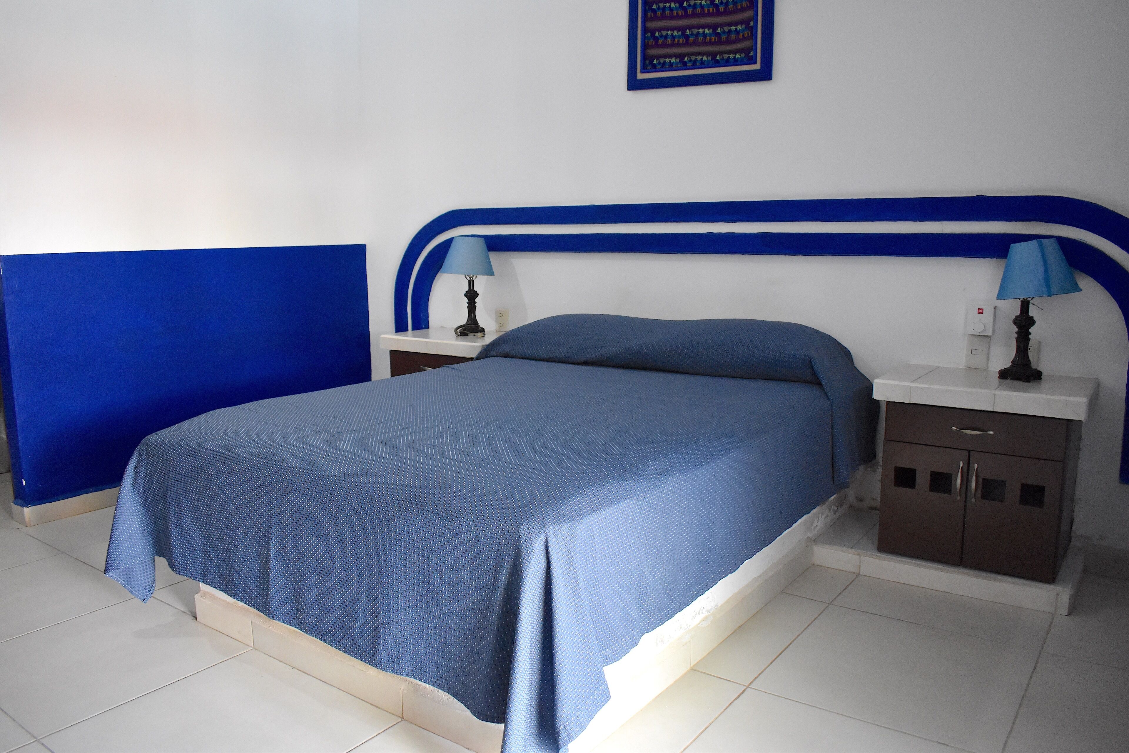 Un lit recouvert d’une couvre-lit bleue, assorti à la tête de lit, flanqué de deux tables de chevet avec des lampes, dans une pièce dont le sol est recouvert de carreaux blancs et dont un mur est peint en bleu.