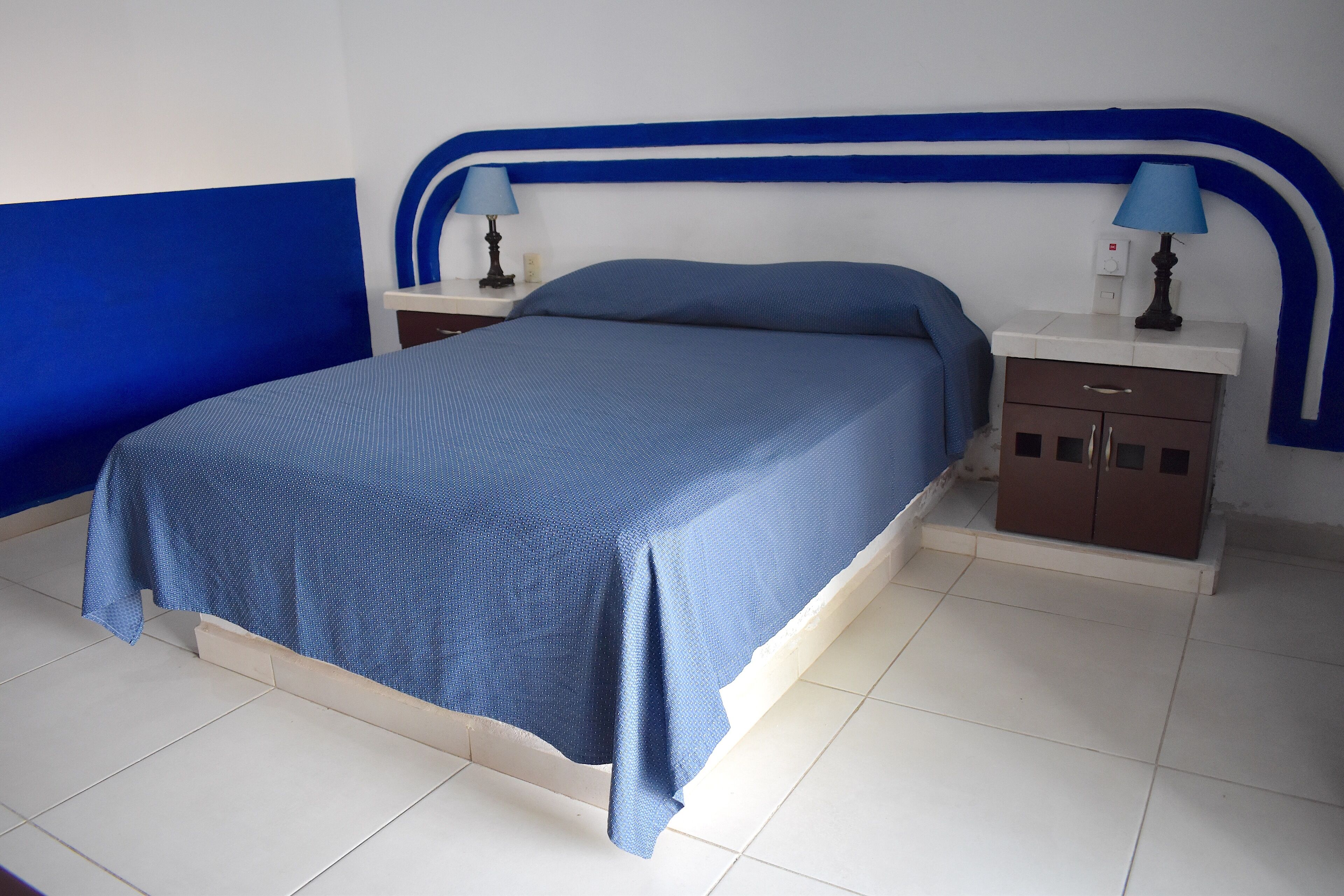 Een bed met een blauwe spreideken, twee nachtkastjes met lampen, en een bruine kast.