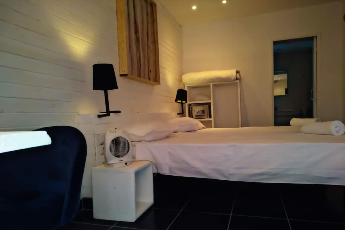 A hotel room with a bed, a small bedside table with a fan, a lamp, and a shelf with a towel.