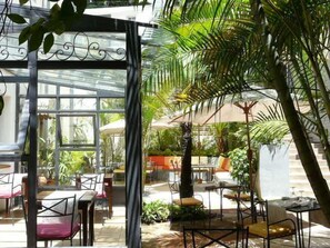Un jardín interior con mesas y sillas, rodeado de abundante vegetación y una estructura de vidrio.