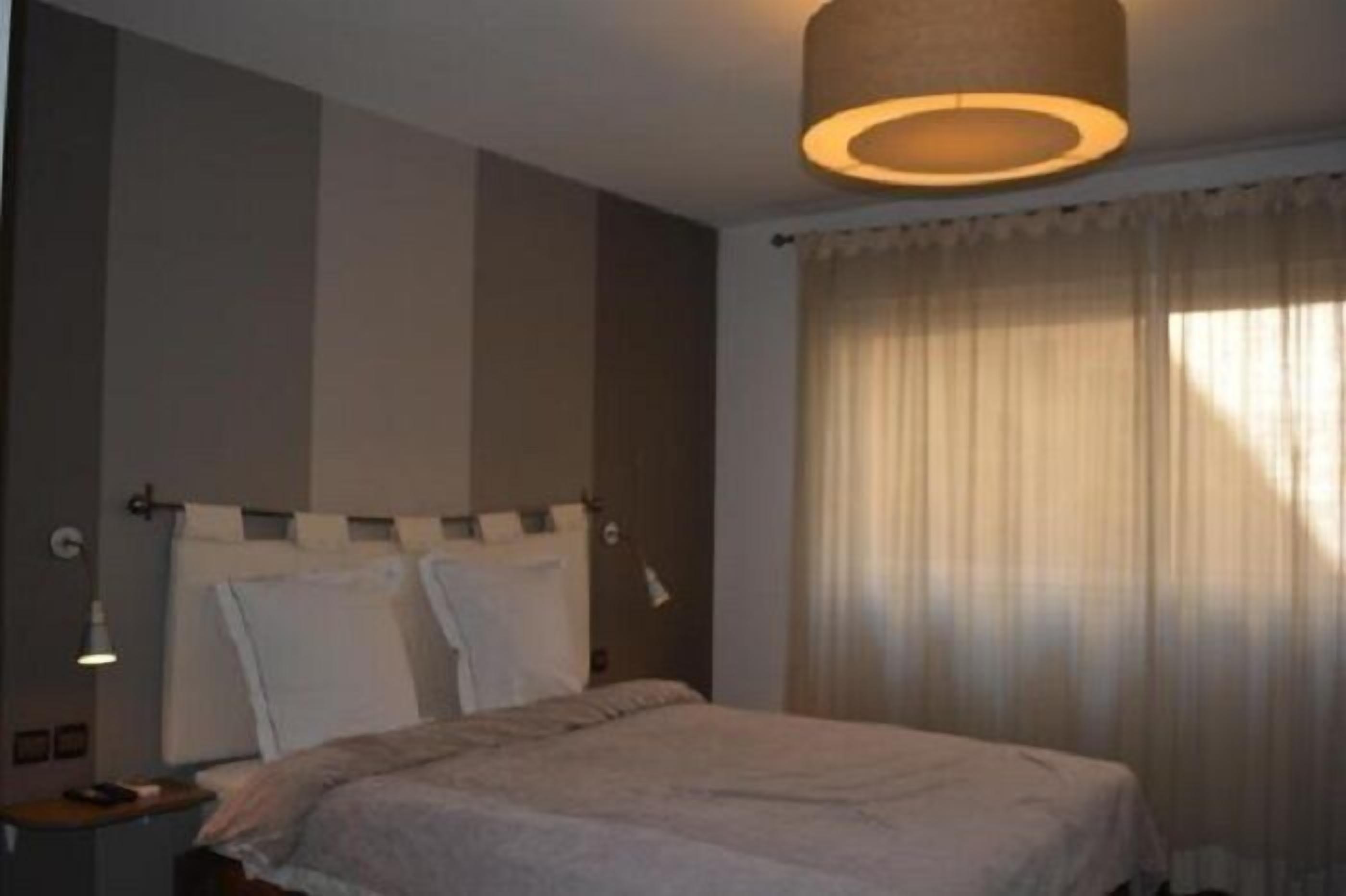 A bedroom with a bed, a bedside lamp, a wall-mounted light, and a window with curtains.