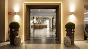 Vestíbulo de hotel con un mostrador de recepción de madera, una mampara de vidrio y dos plantas en macetas con adornos con forma de corazón.
