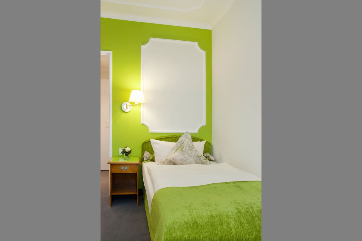Ein Hotelzimmer mit grüner Wand, einem Bett mit grüner Bettdecke, einem Nachttisch mit Lampe und einem Spiegel an der Wand.
