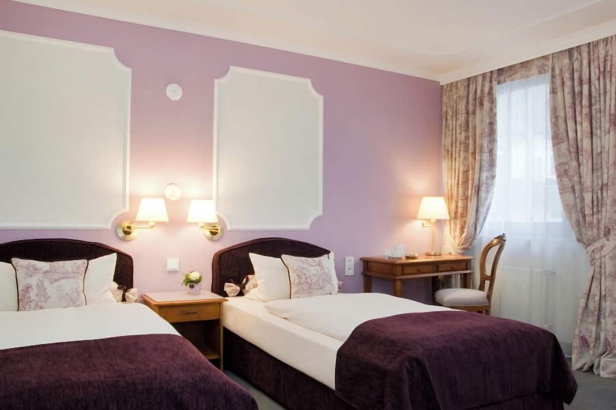 Ein Hotelzimmer mit zwei Betten, einem Schreibtisch und einem Stuhl. An den Wänden sind Lampen angebracht und es gibt ein Fenster mit Vorhängen.