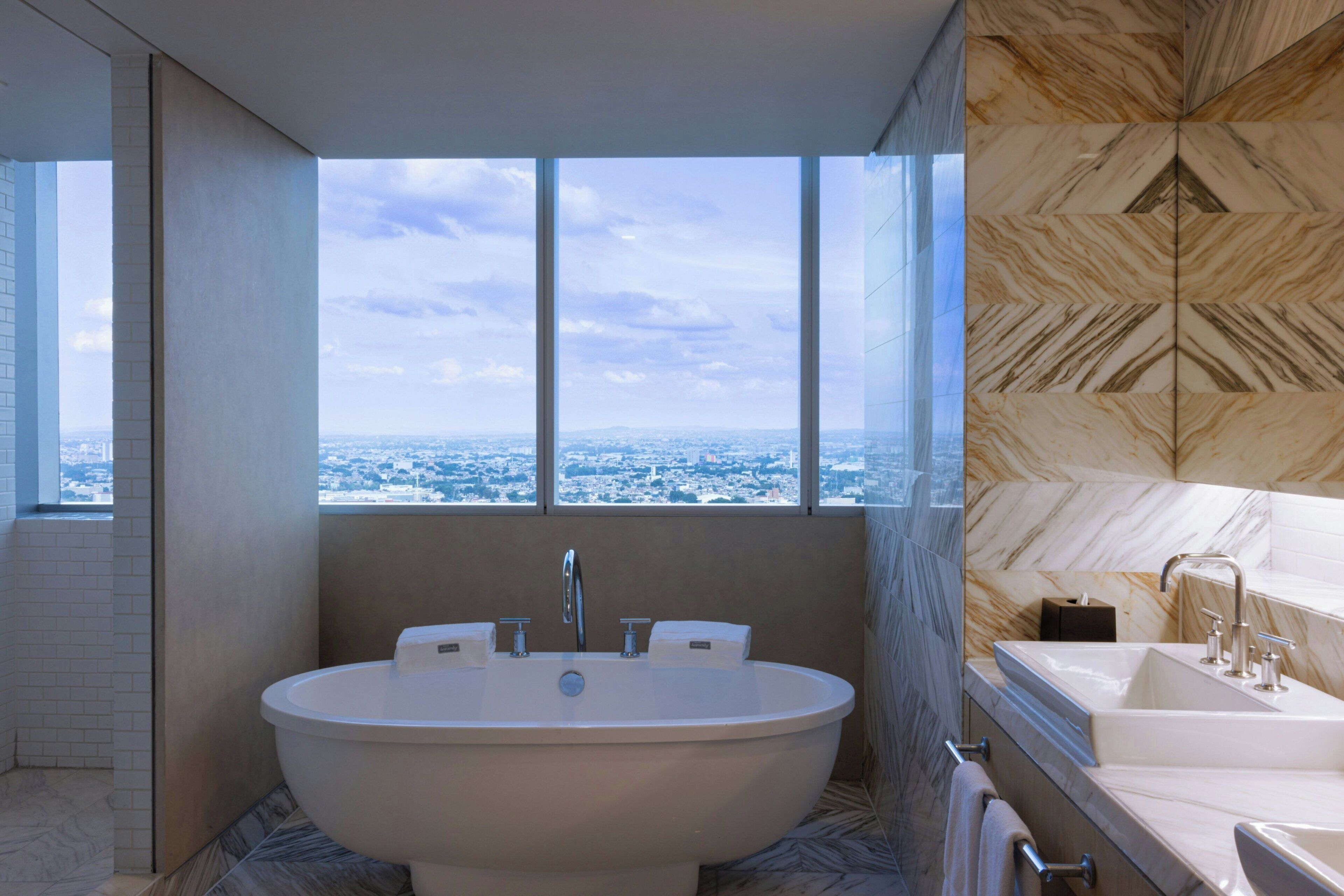Een moderne badkamer met een vrijstaand bad, een grote spiegel en uitzicht op een stadsgezicht door een groot raam.