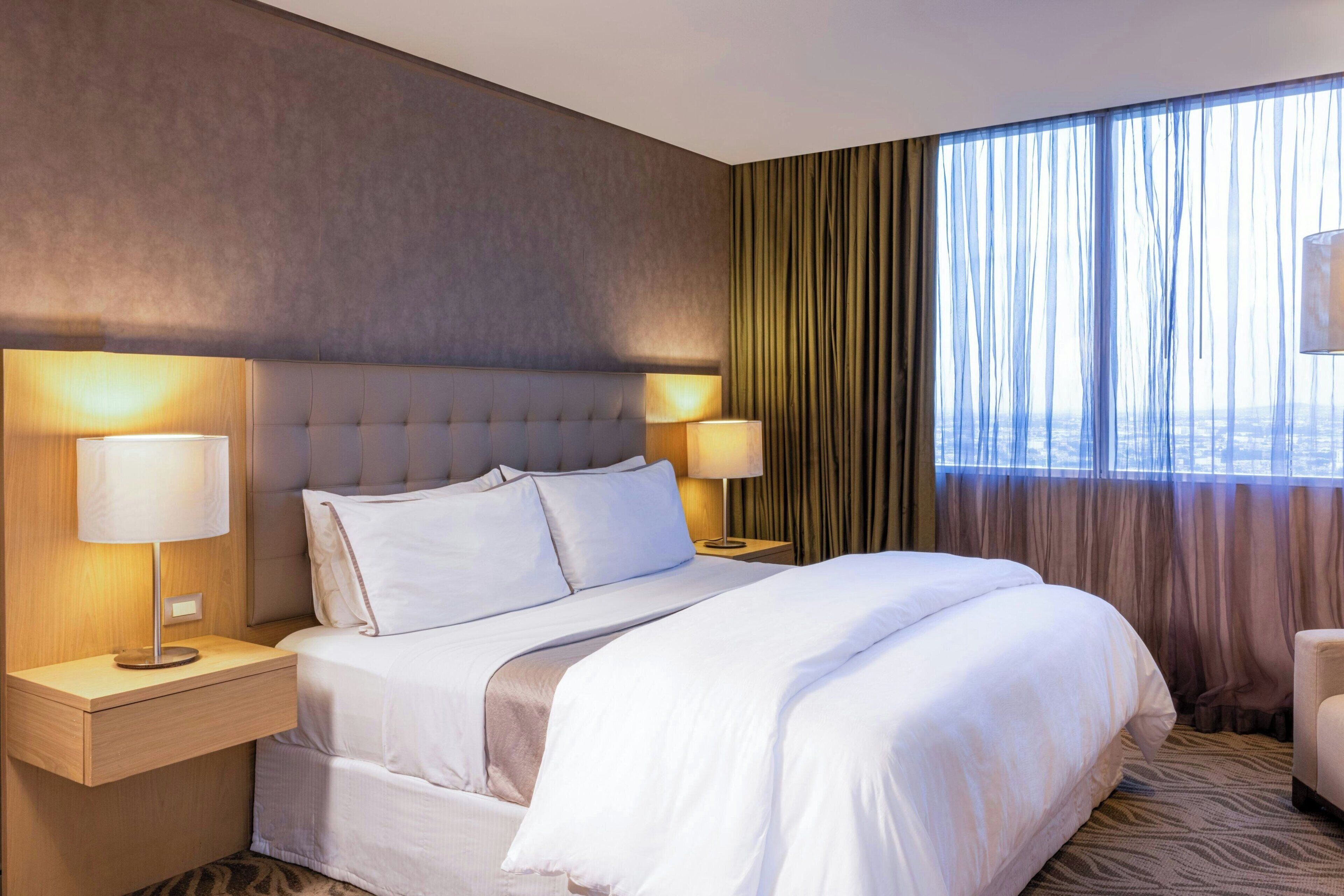 A hotel room with a large bed, two bedside tables with lamps, a view of the city through sheer curtains, and a cityscape visible through the window.