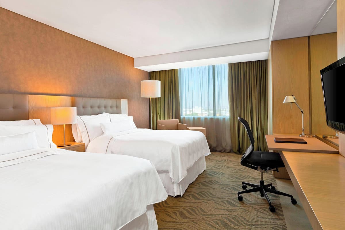 Ein Hotelzimmer mit zwei Betten, einem Schreibtisch, einem Stuhl, einem Fernseher und einem Fenster mit Vorhängen.