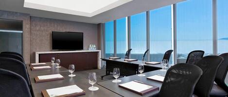 A conference room with a large table, chairs, a flat-screen TV, and a view of the sea through large windows.