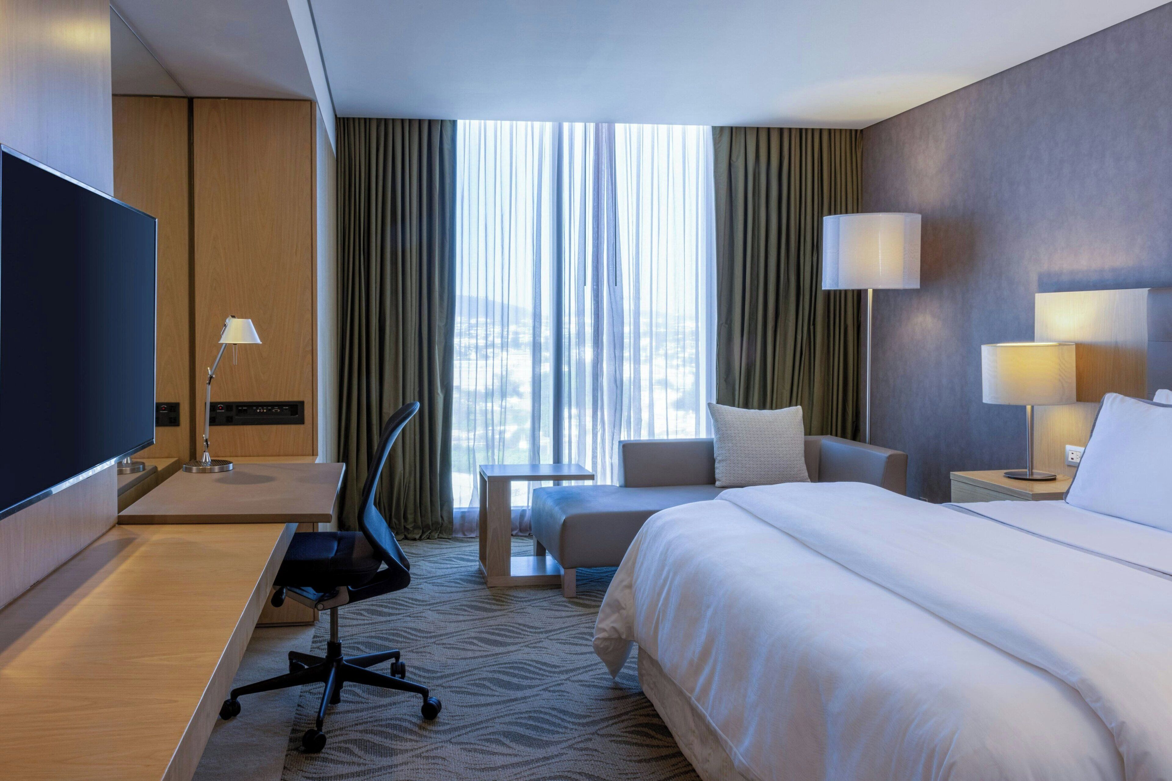 A hotel room with a large bed, a desk with a chair, a television, and a view of the city through the window.