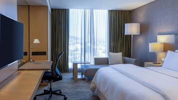 A hotel room with a large bed, a desk with a chair, a television, and a view of the city through the window.