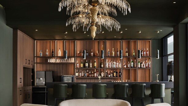 Uma área de bar moderna com prateleiras de madeira exibindo diversas garrafas, um balcão e banquetas de couro.