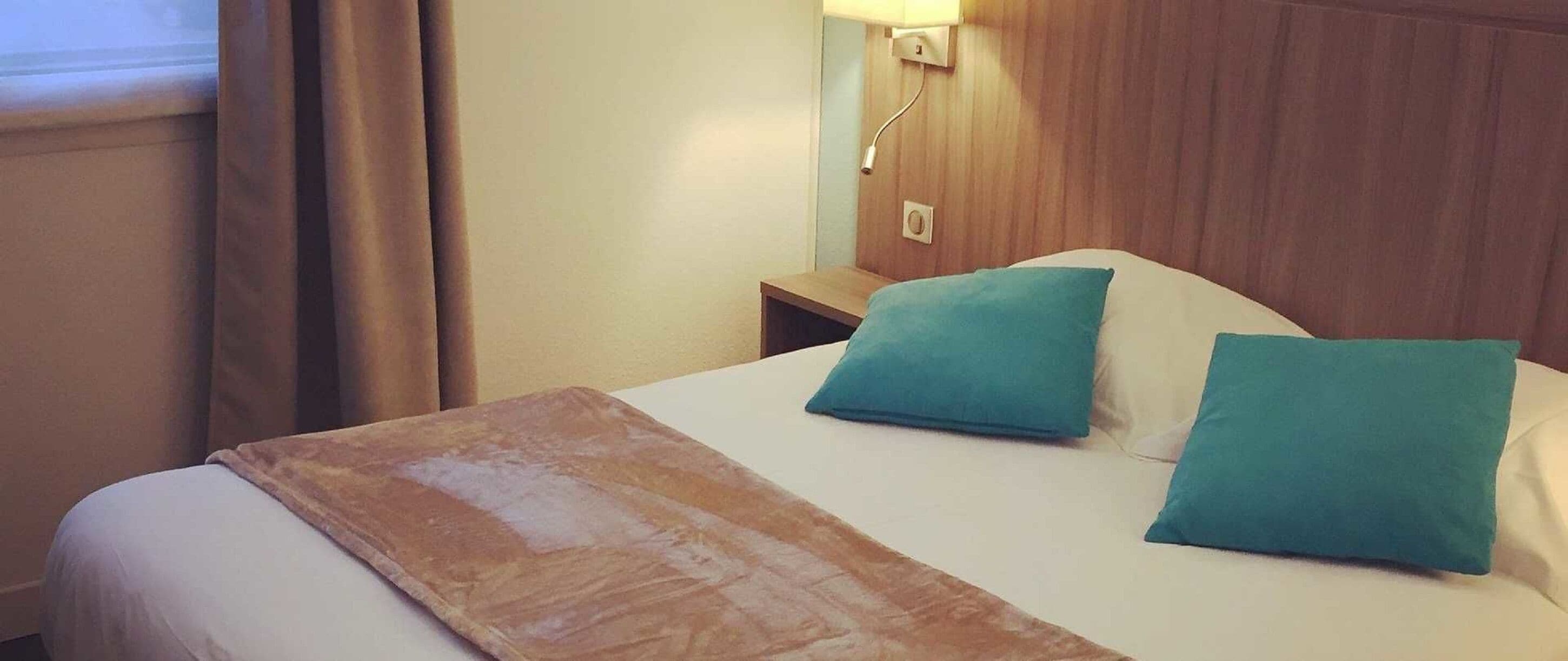 A hotel room with a bed, two pillows, a nightstand, a lamp, and a window with curtains.