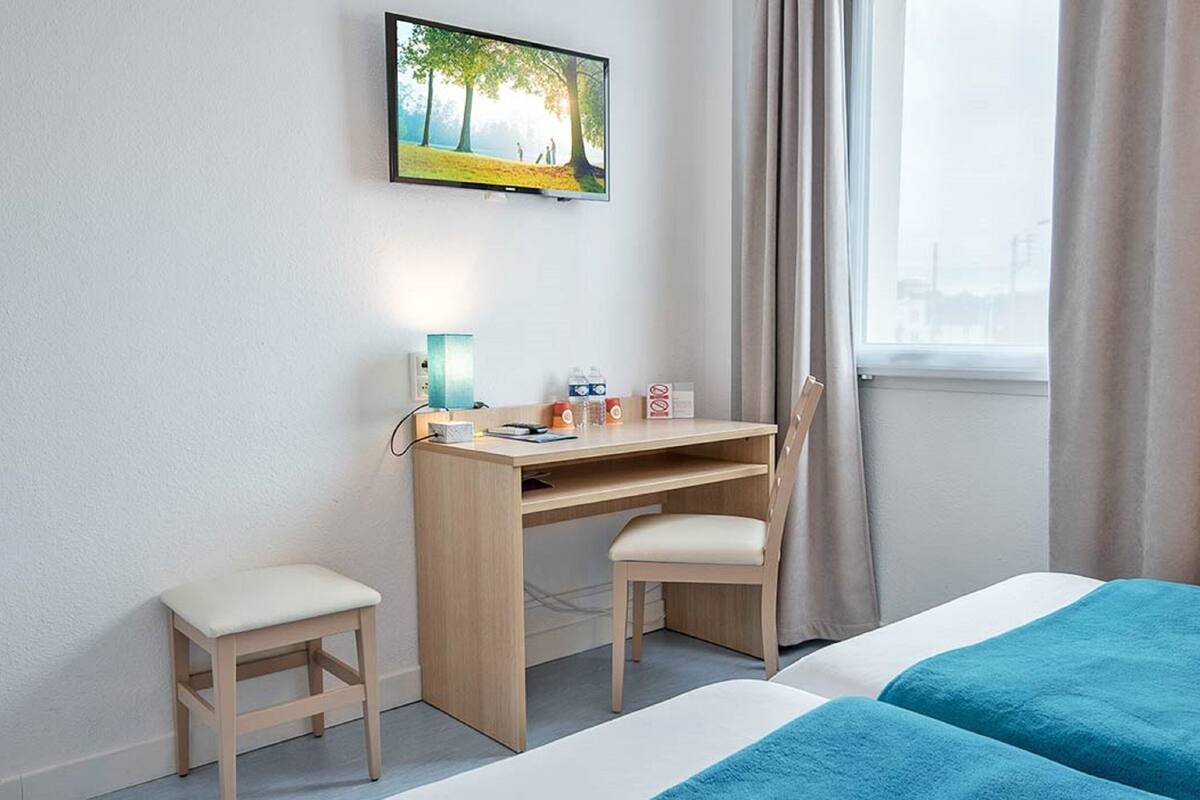 Een hotelkamer met een bed, een bureau met stoel, een aan de muur bevestigde televisie en een raam met gordijnen.